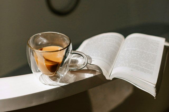 A mug of coffee resting on a bathtub next to a book